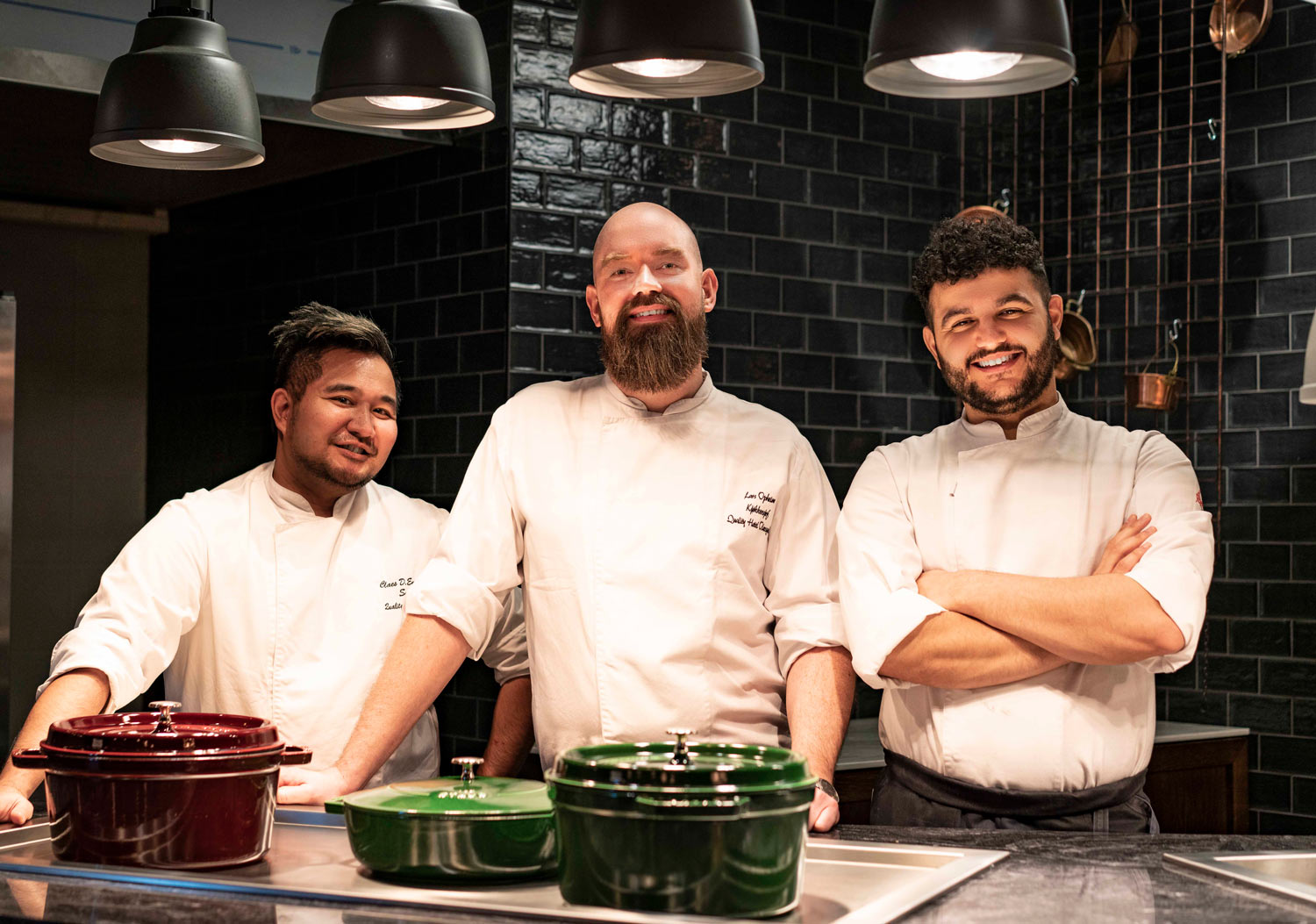 Tre kokker står foran flere gryter og smiler til kameraet.