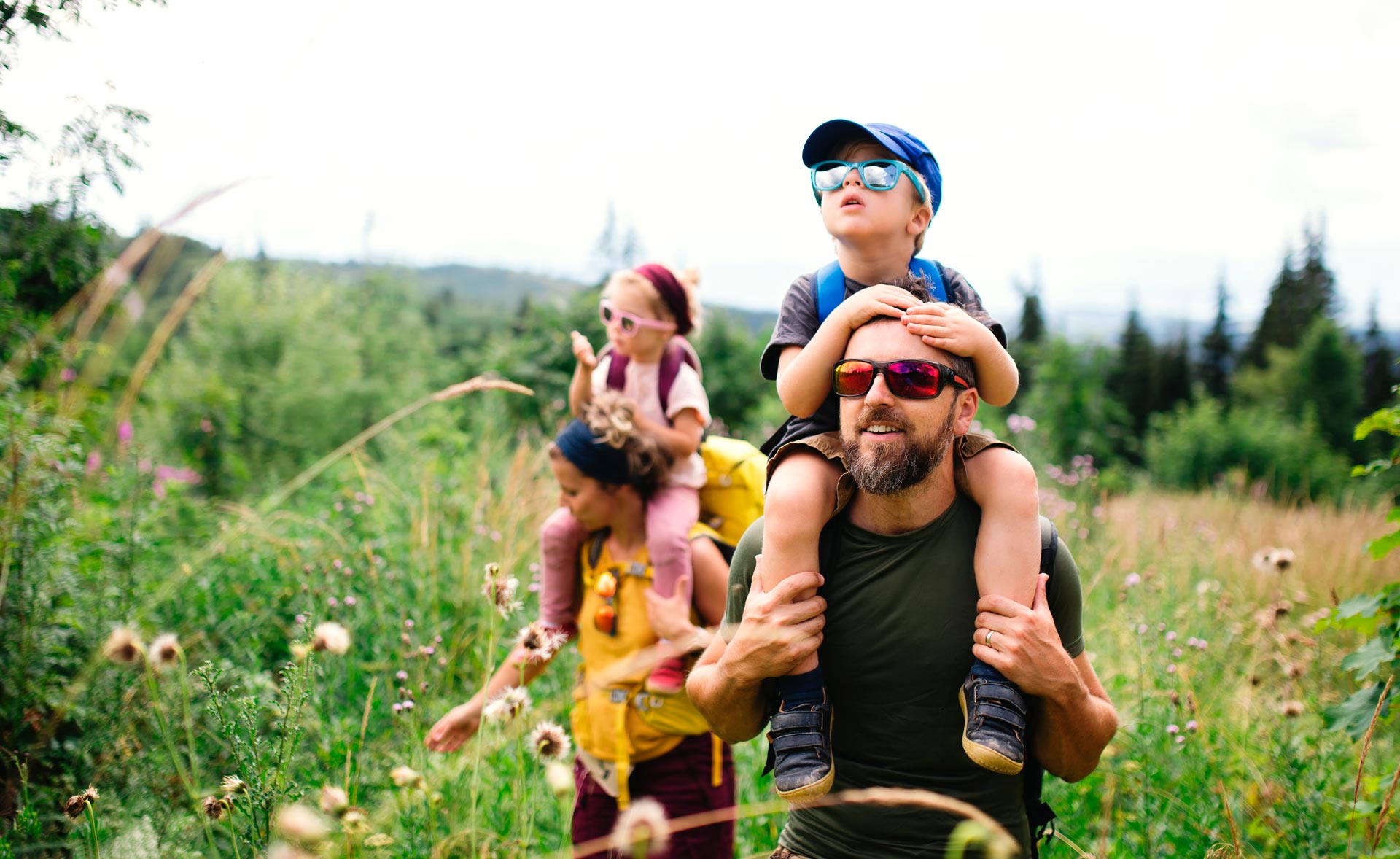 Familie med to voksne og to små barn som sitter på skuldrene til foreldrene, en liten gutt og en liten jente. Familien er på tur i skogen og går i en blomstereng.